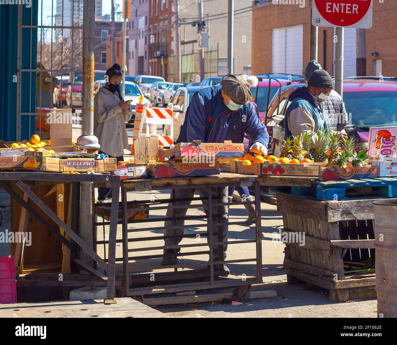 Philadelphia, USA. März 2021, 7th. Eine Frau schaut auf ihr Telefon hinter einem Marktverkäufer im italienischen Markt von Philadelphia, der Produkte zum Verkauf auf einem Tisch organisiert, während zwei Männer an der Straßenecke stehen. Quelle: Kalen Martin/Alamy Live News Stockfoto