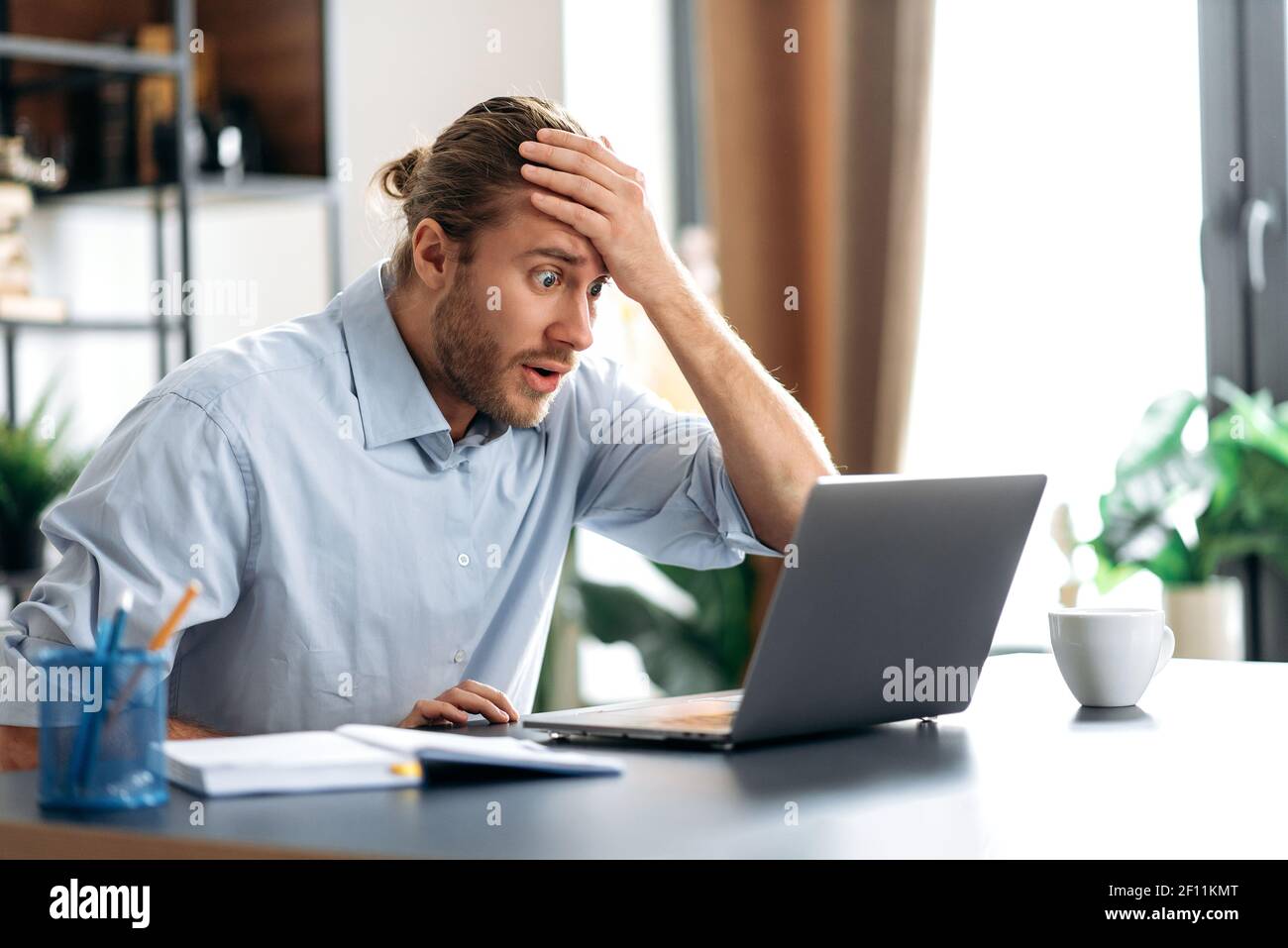 Schockiert verärgert kaukasischen beschäftigt Kerl Blick auf einen Laptop in Verwirrung, erhielt unerwartete Nachrichten, gescheitert einen Deal, schreit, hält seinen Kopf mit der Hand, sitzt am Schreibtisch Stockfoto