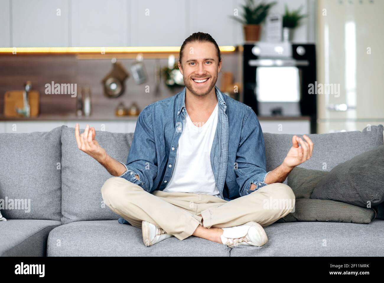 Happy fröhlich kaukasischen stilvollen Kerl in lässiger Kleidung ist entspannend und Meditation während der Sitzung in der Lotus-Position auf dem Sofa zu Hause. Ruhe und Harmonie Konzept Stockfoto