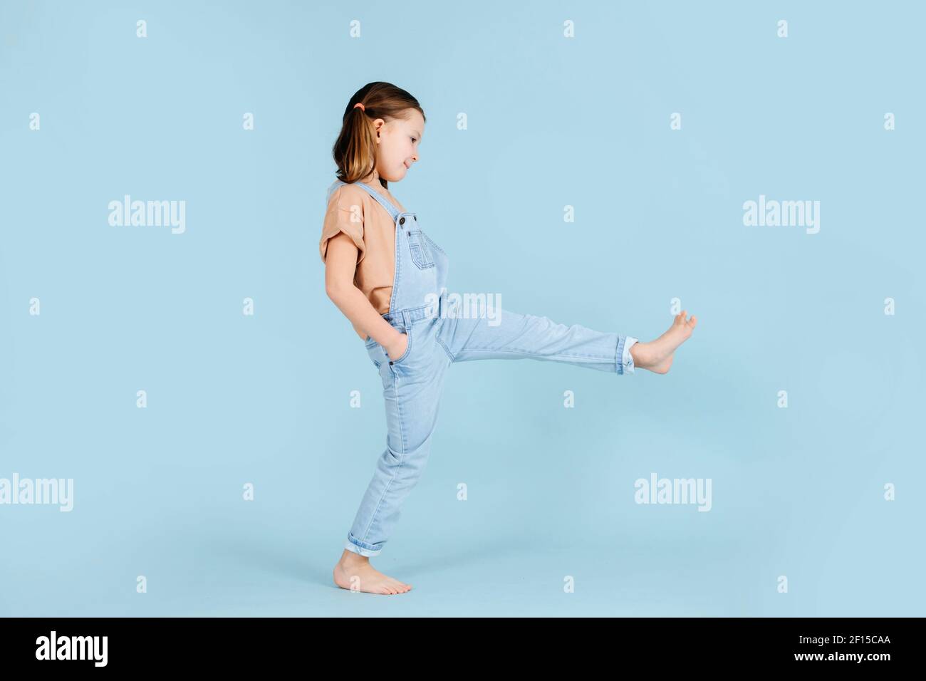 Spielerisch barfuß kleines Mädchen tun breite Schritte mit geraden Beinen über blauen Hintergrund.. Sie hat zwei Pferdeschwänze, trägt Overalls. Hände in den Taschen. Stockfoto