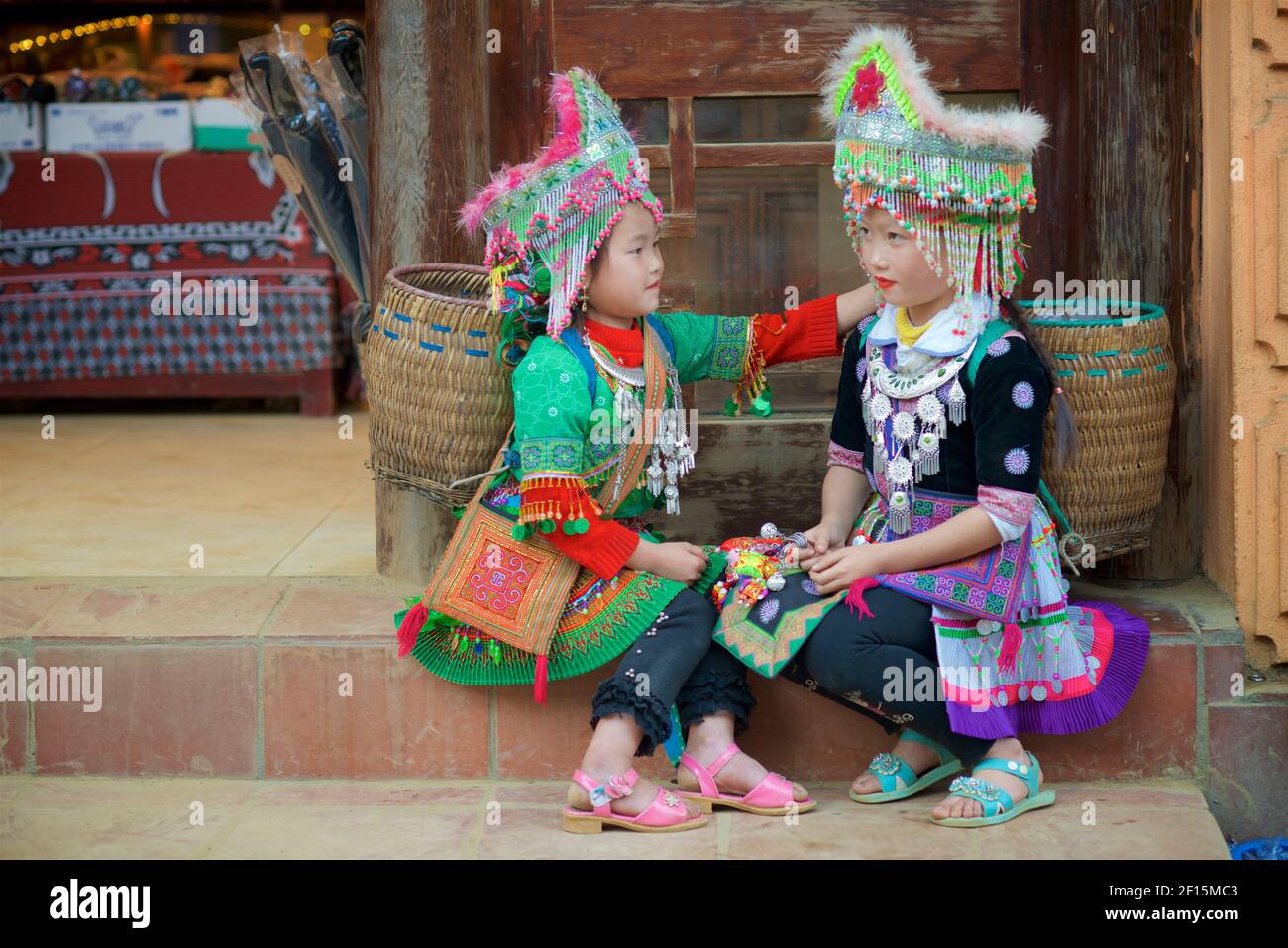 Zwei Blumen-Hmong-Mädchen mit Kopfbedeckungen sitzen auf einer Stufe und tragen indigene Hügelstämme der Provinz Lao Cai, Vietnam. Sapa Stockfoto