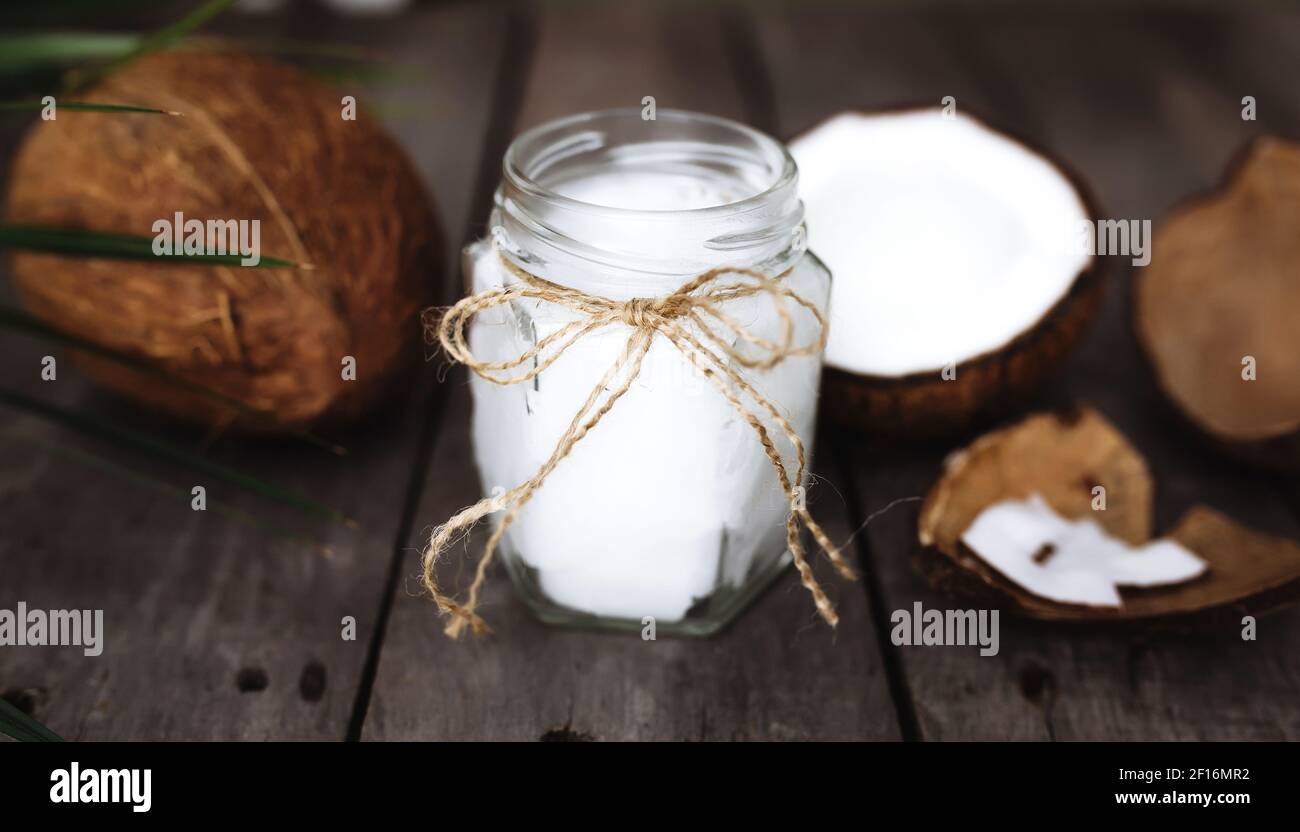 Gebrochene Kokosnüsse auf grauem Holzhintergrund mit Glas rohen Bio-Kokosnussöl extra vergine und Palmblatt. Weißes Kokosnusspulpe. Hochwertige Fotos Stockfoto