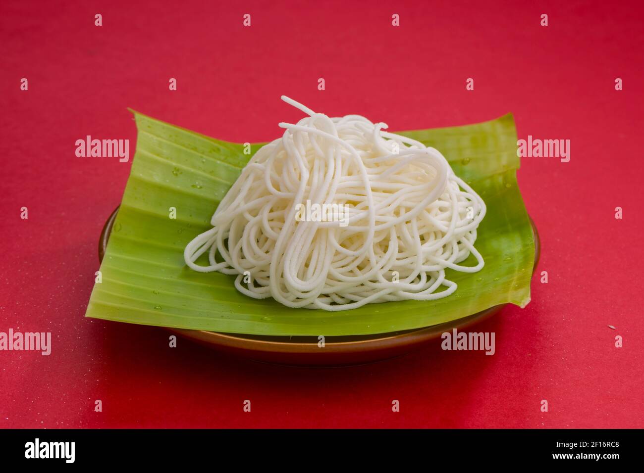 Ideyappam oder Noolappam in einem frischen grünen Bananenblatt mit rotem Strukturhintergrund angeordnet. Stockfoto