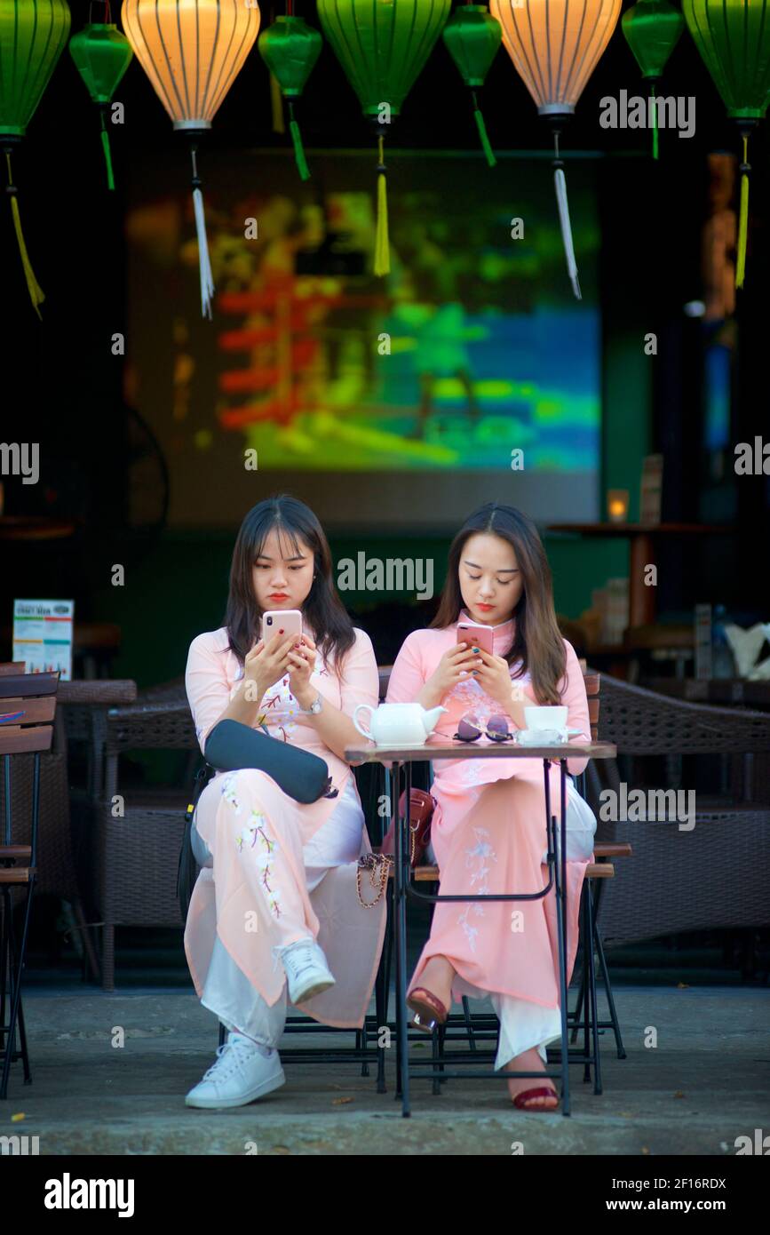 Asiatische Frauen tragen vietnamesischen Stil Ao Dai und mit ihren Mobiltelefonen in einem Straßencafé in Hoi an, Vietnam Stockfoto