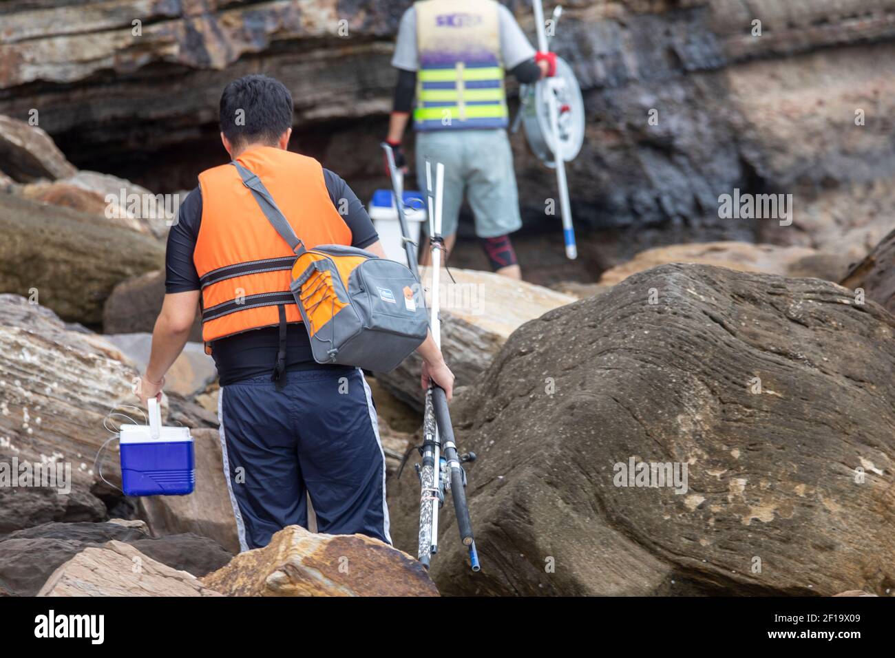 Zwei asiatische Männer mit Angelausrüstung und Ruten fahren über die Felsen zu einem Angelplatz in Sydney, Australien Stockfoto