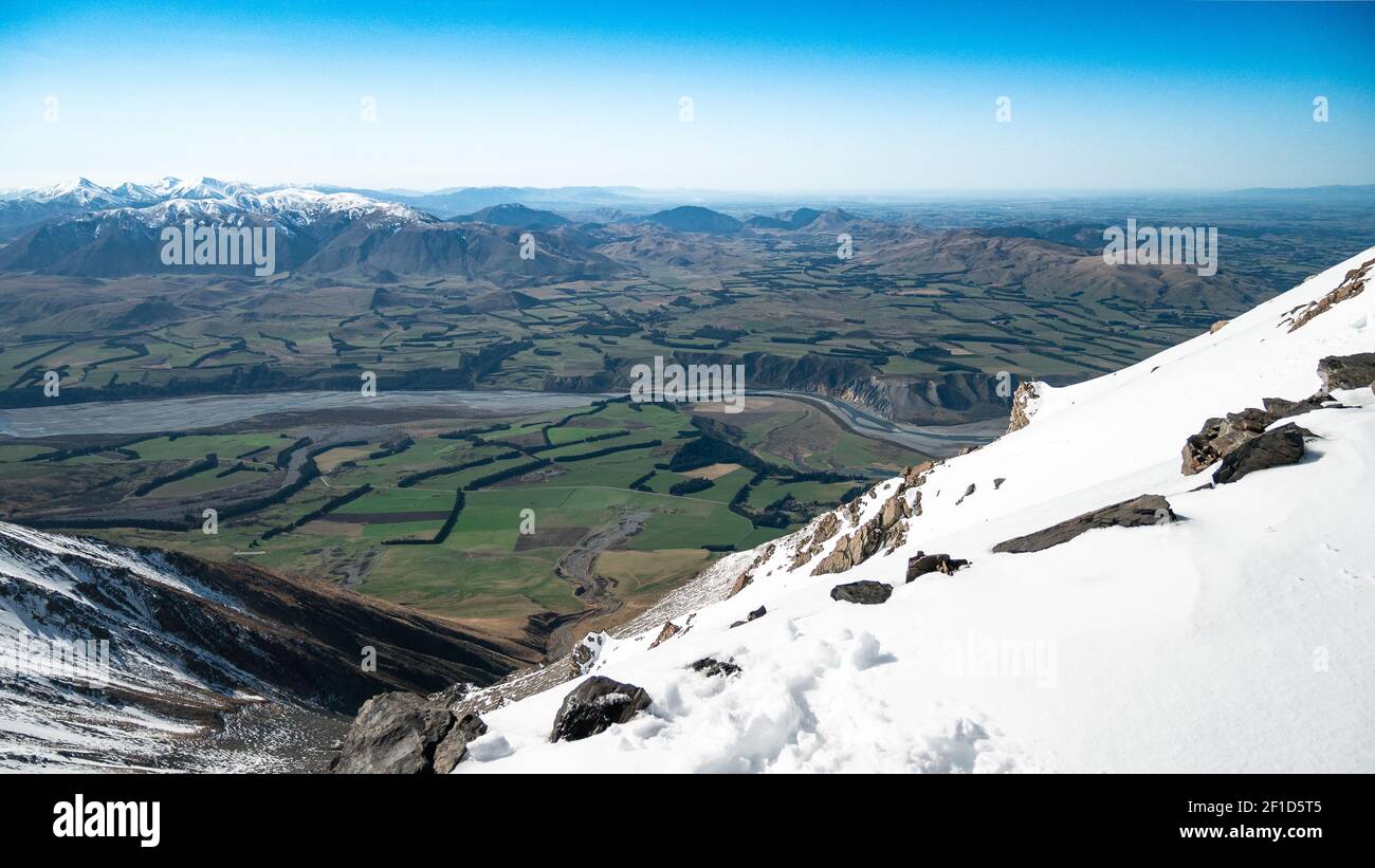 Schöne Aussicht von der Spitze des verschneiten Berges hinunter ins grüne Tal mit Bergkulisse und blauem Himmel. Aufgenommen in Mount Hutt, Canterbury Region, Neuseeland Stockfoto