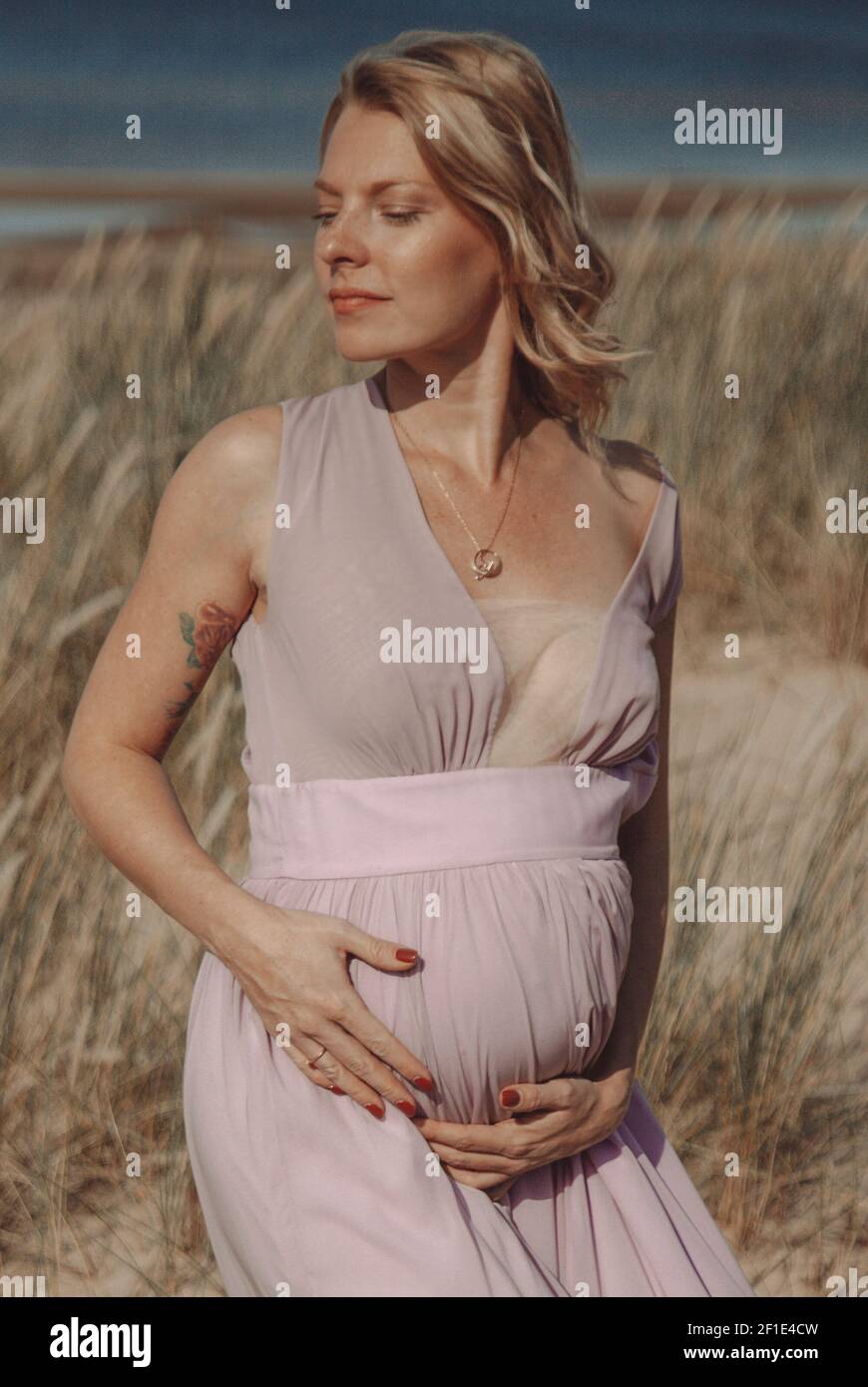Junge, blonde Schwangerin trägt am Strand ein weißes Spitzenkleid, das den Bauch der Schwangerschaft hält. Freier Speicherplatz. Mutterschaft. Stockfoto