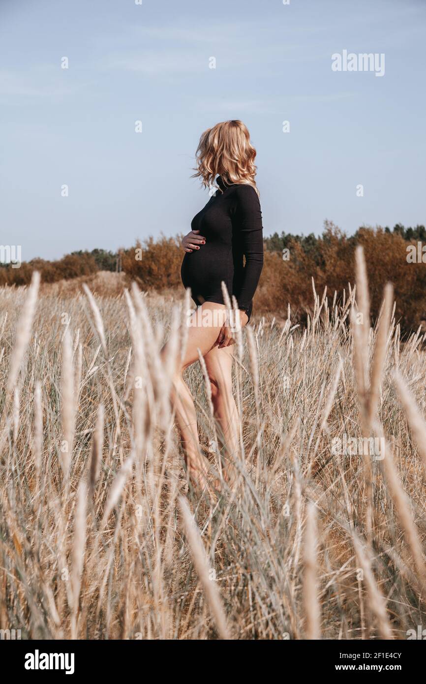Junge, blonde Schwangerin am Strand gebogenes Gras trägt schwarzen Body und hält den Bauch der Schwangerschaft. Freier Speicherplatz. Mutterschaft. Konzept der Sommerschwanger. Stockfoto