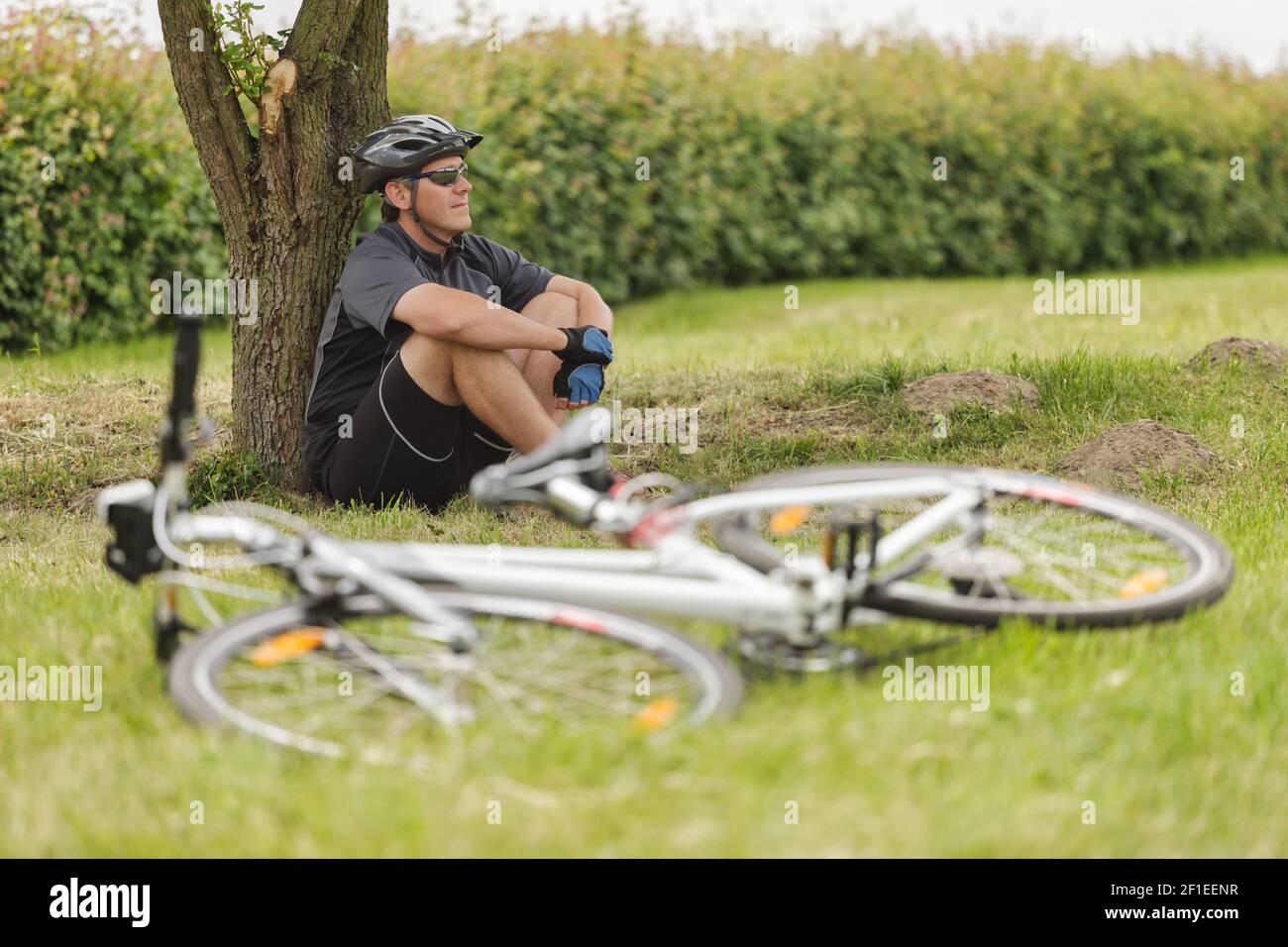Radfahrer ruht auf einer Wiese unter einem Baum, unscharfes Fahrrad im Vordergrund sichtbar Stockfoto