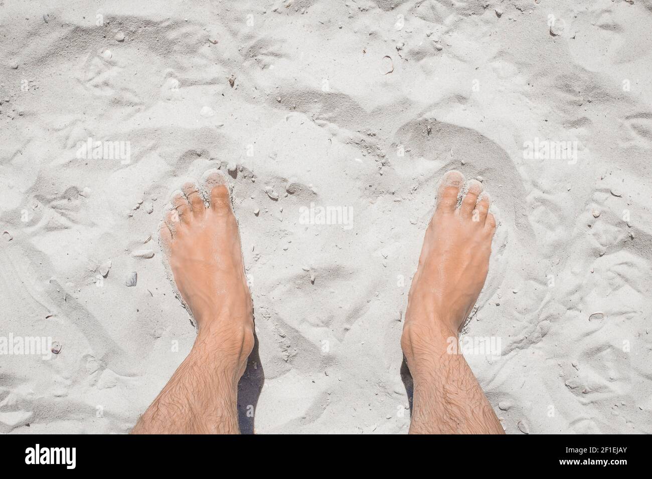 Die Beine des jungen Mannes stehen entspannt auf dem weißen Strandsand, der Blick von oben. Stockfoto