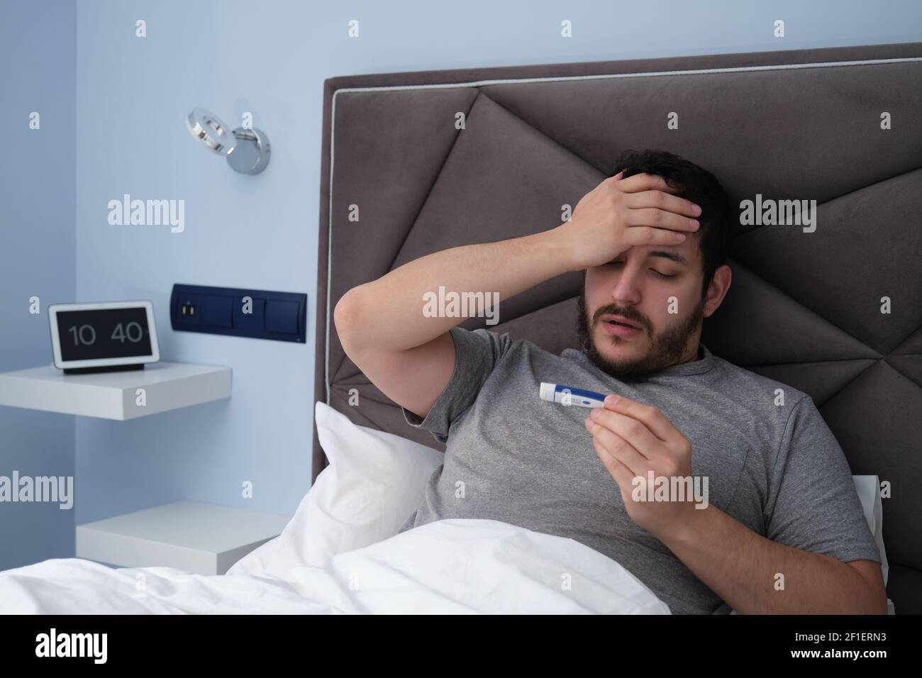 Junger latein krank im Bett mit einem Thermometer in der Hand. Digitale Uhr im Hintergrund. Stockfoto