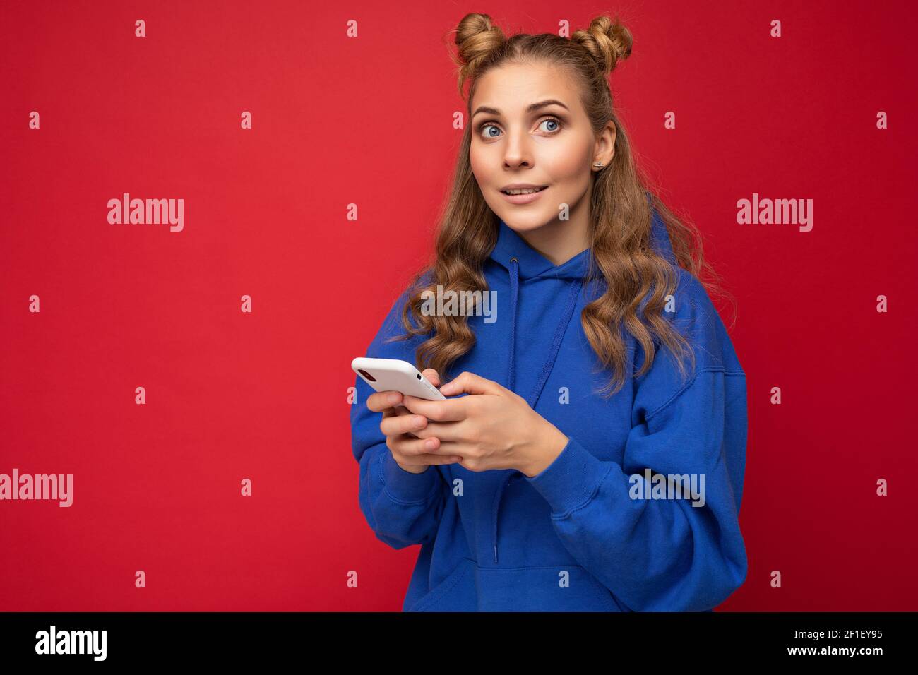 Attraktive junge blonde Frau trägt blau stilvolle Hoodie stehend isoliert Über rotem Hintergrund SMS-Nachricht über Telefon mit Blick auf die Seite Stockfoto