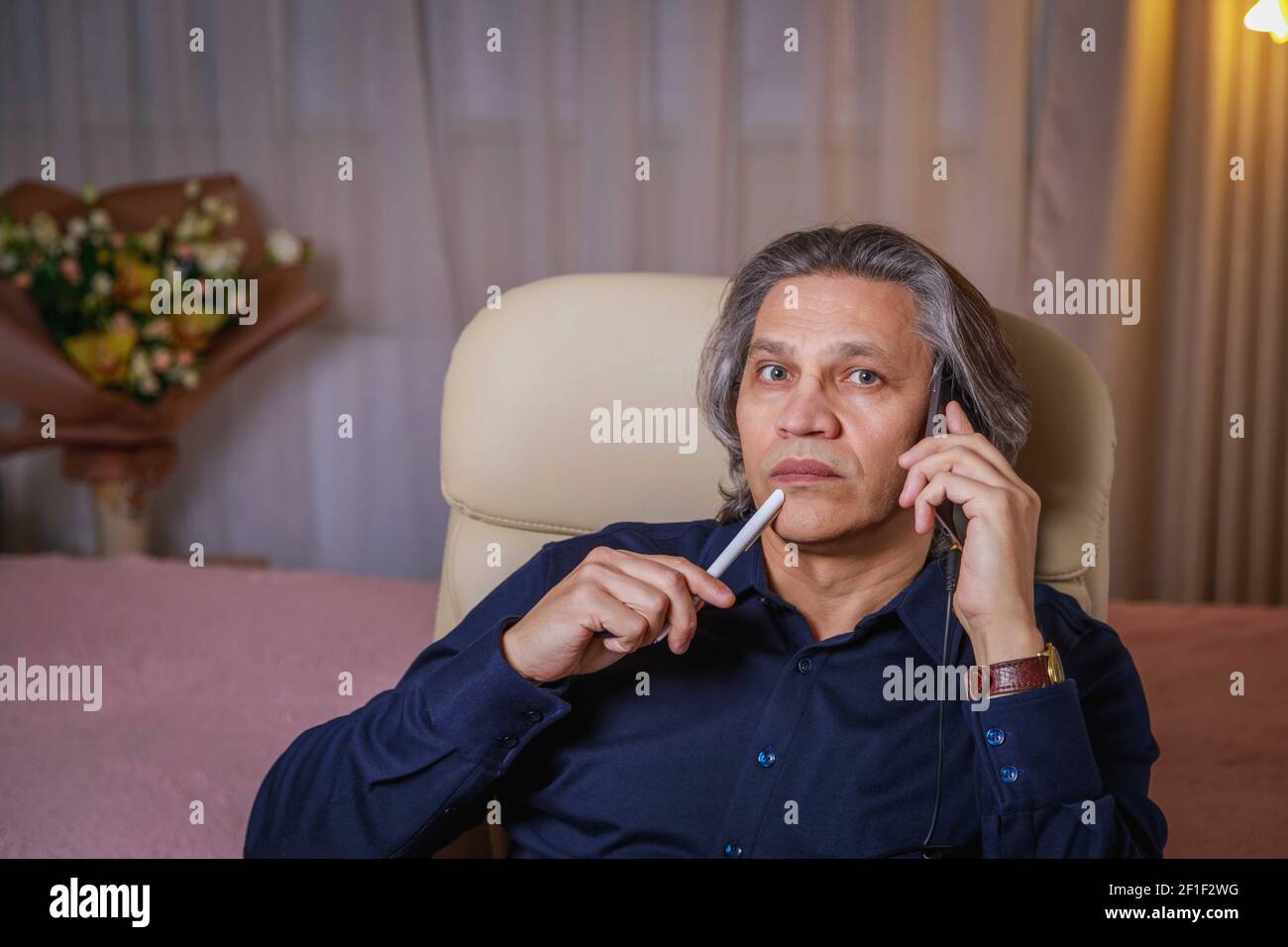 Ein 50-jähriger Mann mit langen Haaren spricht zu Hause auf einem Stuhl am Telefon. Erholsame Freude. Stockfoto