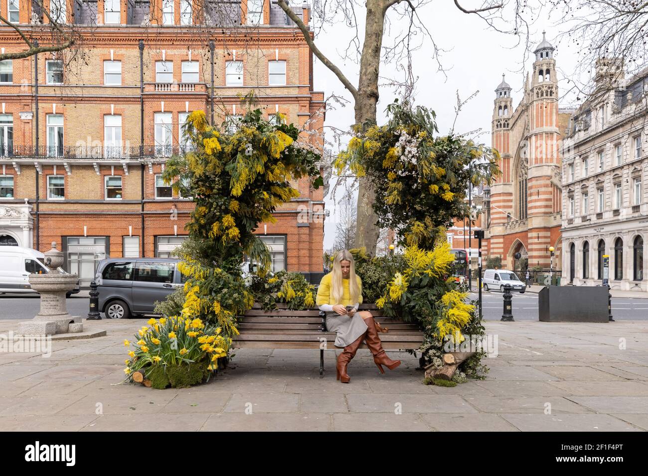 Ein weibliches Modell posiert für Fotos an einer mit Blumen dekorierten Bank.London Event Florist, Warren Bushaway, feiert den Internationalen Frauentag mit einer Banksy-Stil Blumenarrangement in Sloane Square. Stockfoto