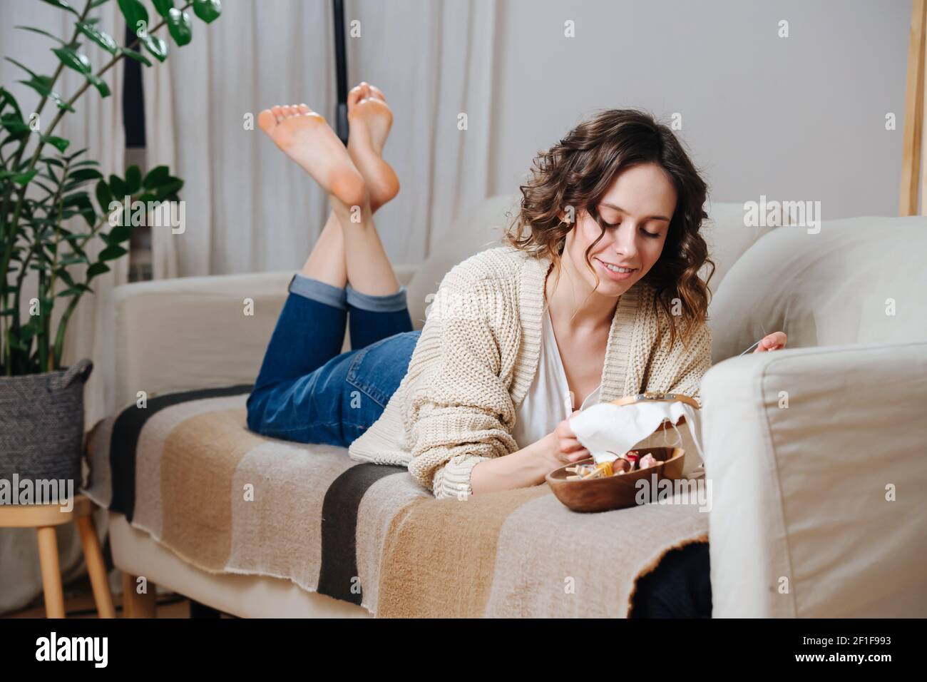 Glückliche junge Frau Nähen in Komfort ihrer Wohnung. Sie liegt auf einer Couch und stickt ein buntes Bild auf einer Schlaufe. Stockfoto