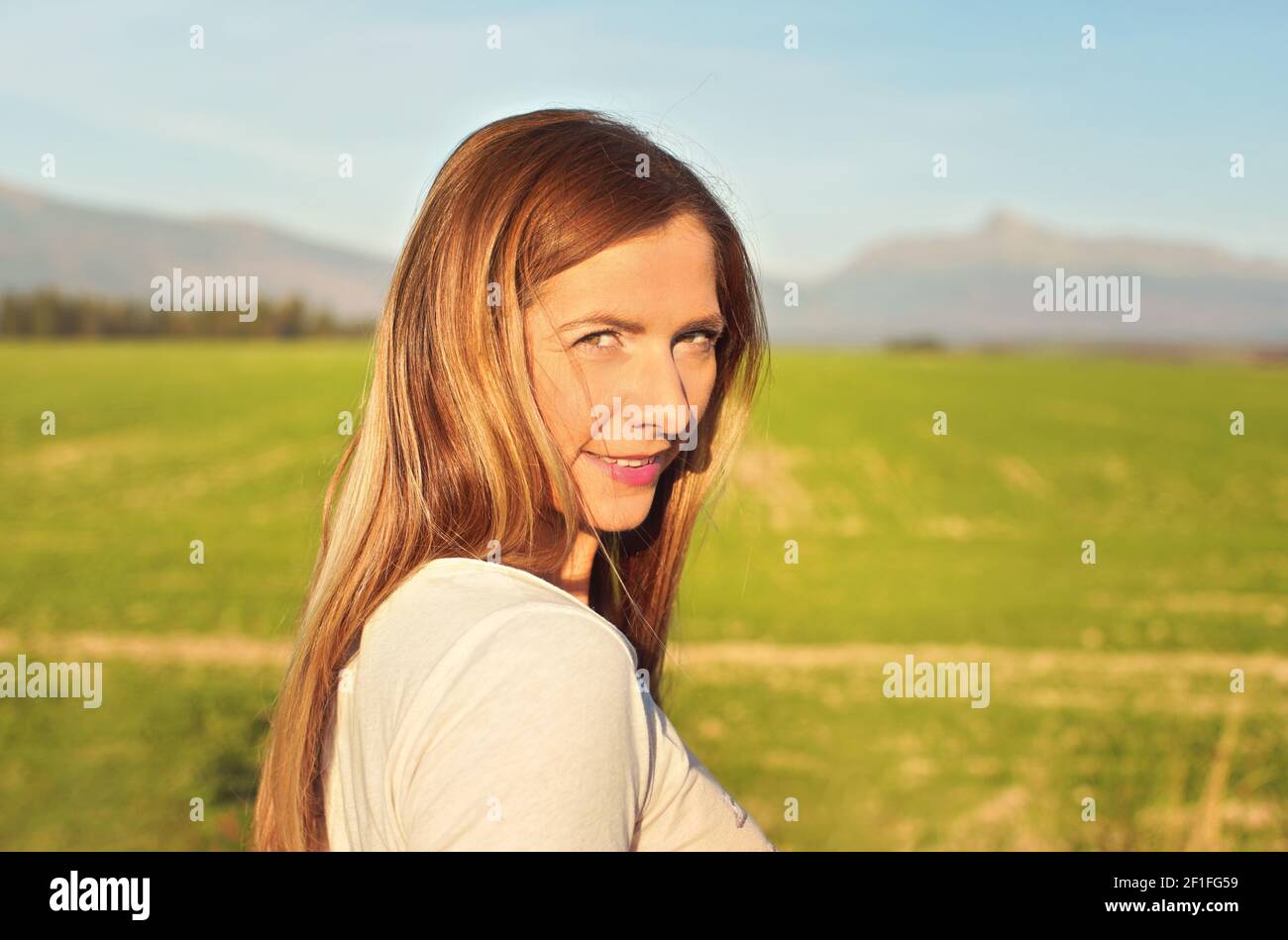 Porträt einer jungen Frau, beleuchtet von der Nachmittagssonne, verschwommene Felder und Berge im Hintergrund Stockfoto