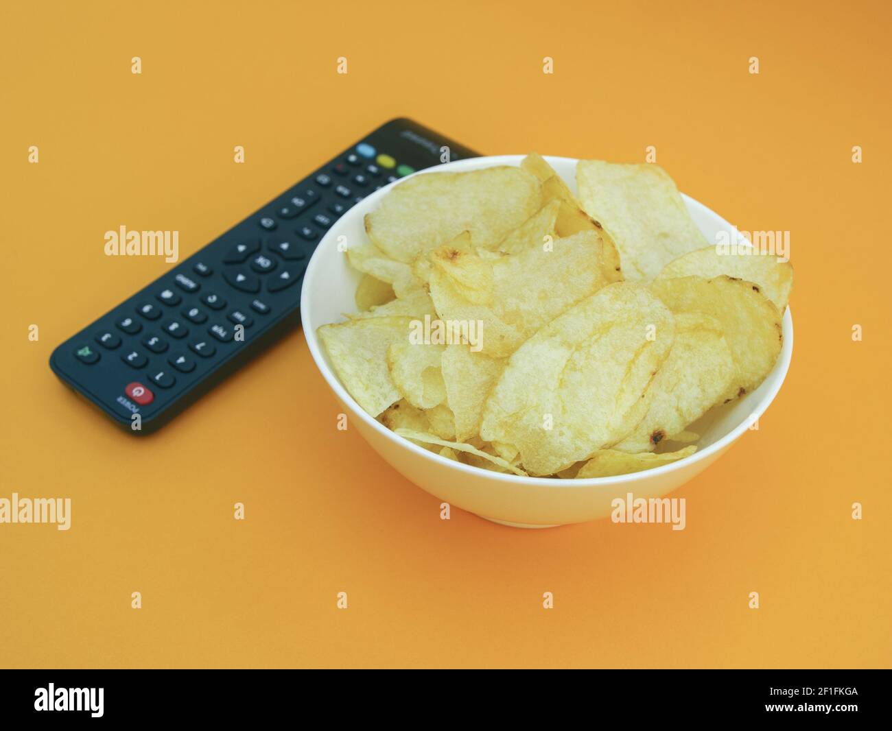 Chips in der Schüssel und TV-Fernbedienung isoliert auf orangefarbenem Hintergrund, Foto Stockfoto