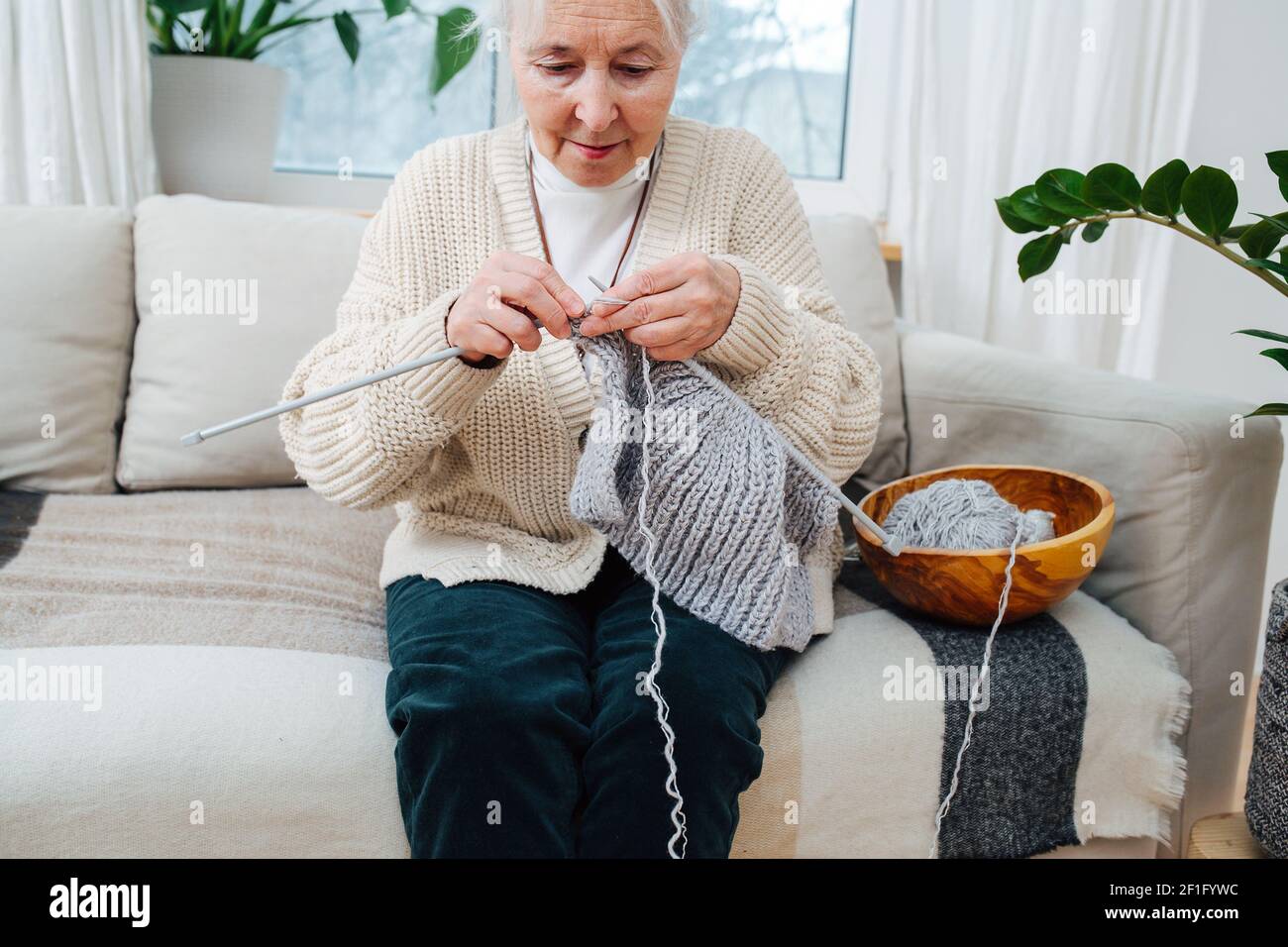 Eine ältere Frau von 70 Jahren stickt mit Stricknadeln, während sie auf dem Sofa sitzt. Stockfoto