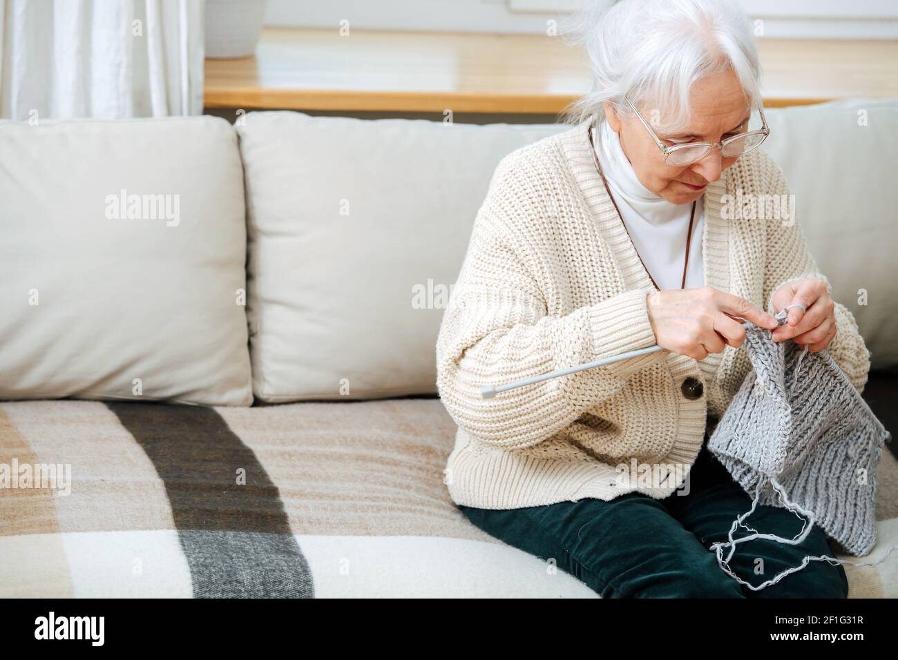 Nahaufnahme einer älteren Frau, die sich mit dem Stricken auf dem Sofa zu Hause beschäftigt. Sie hat graues Haar und Hyperopie-Brille Stockfoto