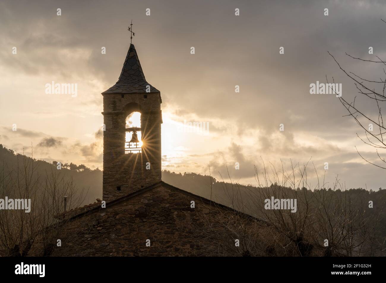 Eine massive Bergkirche aus Stein mit Kirchturm und Sonne Stern, der im Winter unter einem bewölkten Himmel durchscheint Abend Stockfoto