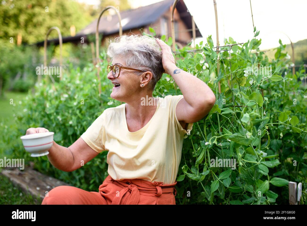 Porträt einer älteren Frau, die im Freien am Gemüsegarten sitzt und eine Tasse Kaffee hält. Stockfoto