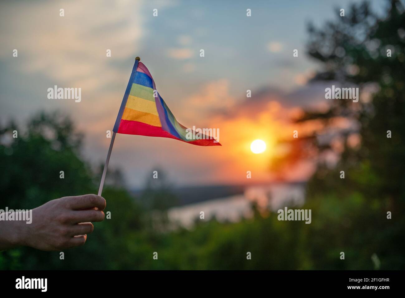 Eine Hand schwingt bei Sonnenuntergang eine bunte Homosexuell Stolz LGBT Regenbogenfahne auf einer natürlichen Landschaft im Sommer. Stockfoto