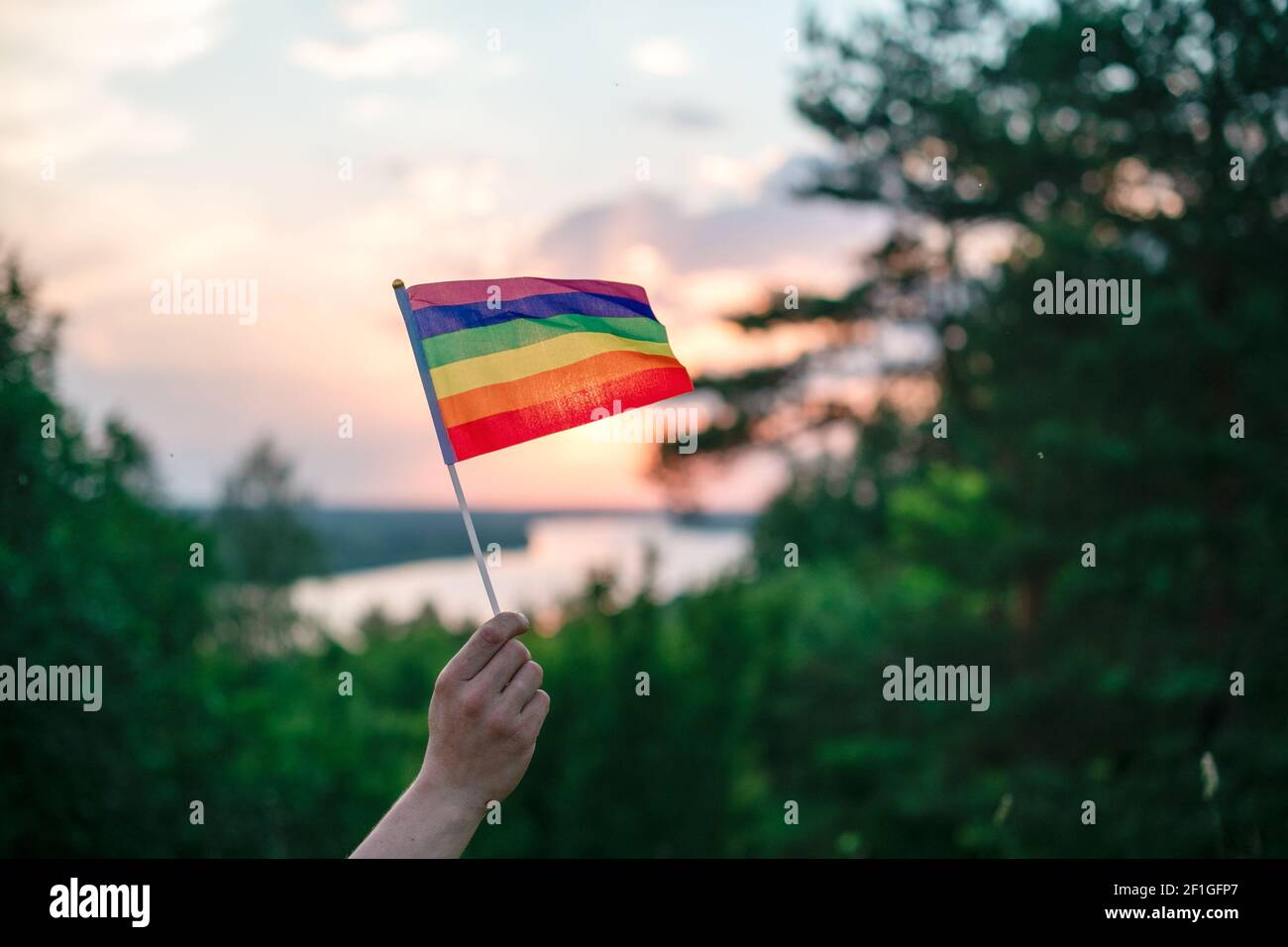 Eine Hand schwingt bei Sonnenuntergang eine bunte Homosexuell Stolz LGBT Regenbogenfahne auf einer natürlichen Landschaft im Sommer. Stockfoto