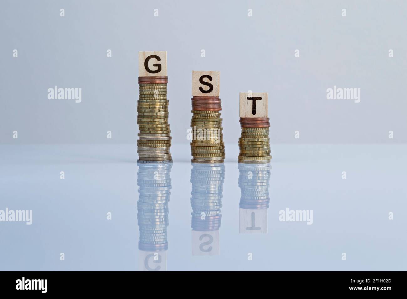 Wort „GST“ auf Holzblöcken auf absteigenden Münzstapeln auf Grau. Konzeptfoto der Reduzierung von Mehrwertsteuer, Wirtschaft, Unternehmen und Finanzen Stockfoto