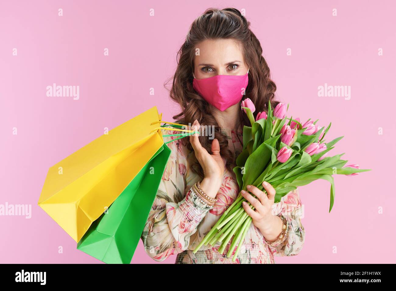Moderne Frau im floralen Kleid mit Tulpen Bouquet, rosa Maske und Einkaufstaschen isoliert auf rosa. Stockfoto