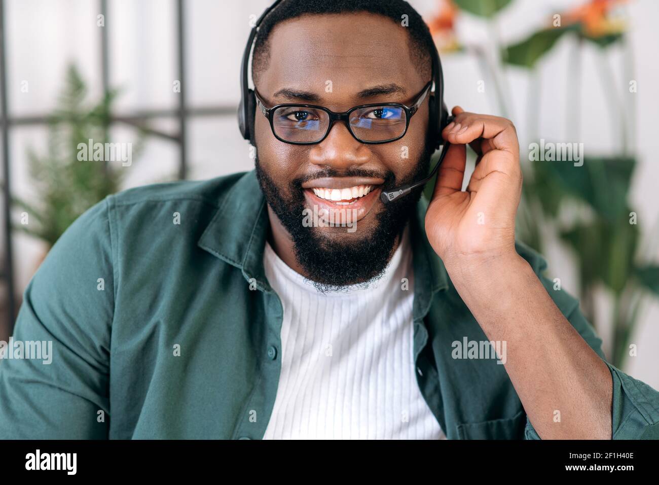 Nahaufnahme eines attraktiven, selbstbewussten bärtigen afroamerikanischen Callcenter-Betreibers oder Geschäftsführers. Schwarzer Geschäftsmann in Headset und stylish Shirt sieht auf die Kamera und freundlich Stockfoto