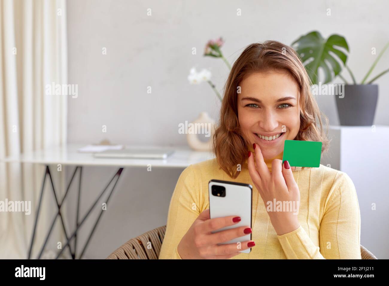 Entzückt weiblich mit Plastikkarte und Smartphone macht Online-Kauf Während Sie die Kamera zu Hause betrachten Stockfoto
