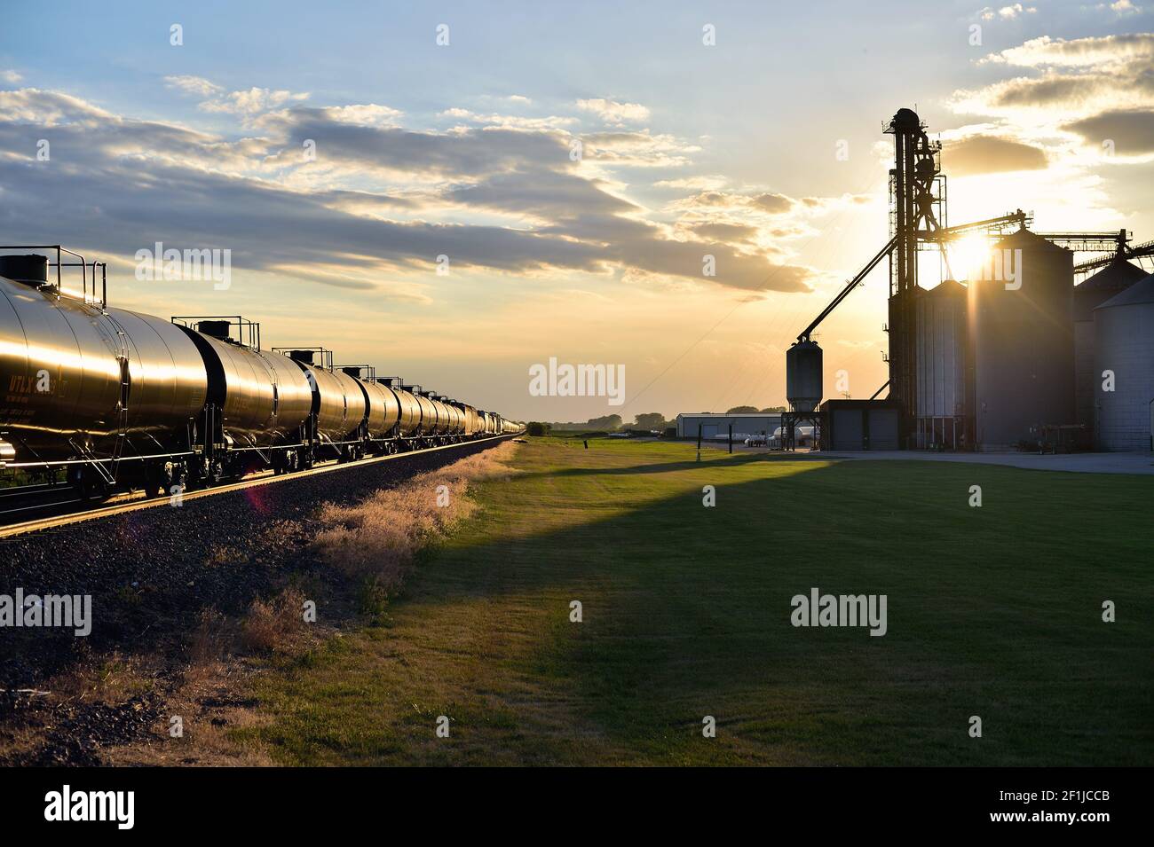 Maple Park, Illinois, USA. Ein Güterzug der Union Pacific spiegelt die untergehende Sonne wider, wenn er an einer landwirtschaftlichen Genossenschaft vorbei nach Westen fährt. Stockfoto