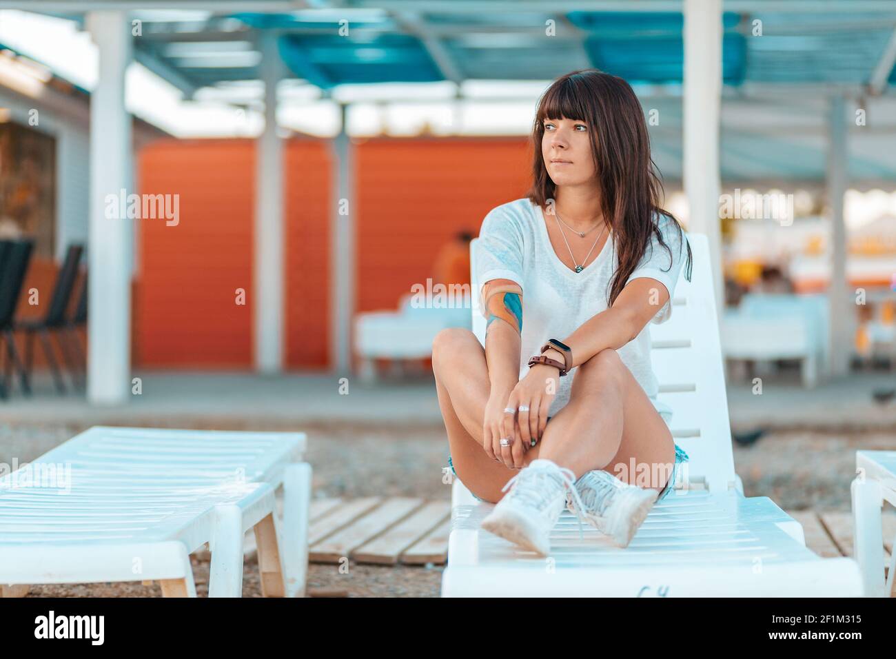 Gebräunte hübsche Frau mit Tattoos, die auf einer Chaiselongue sitzt. Cafés am Wasser und der Strand. Das Konzept des Sommerurlaubs. Stockfoto