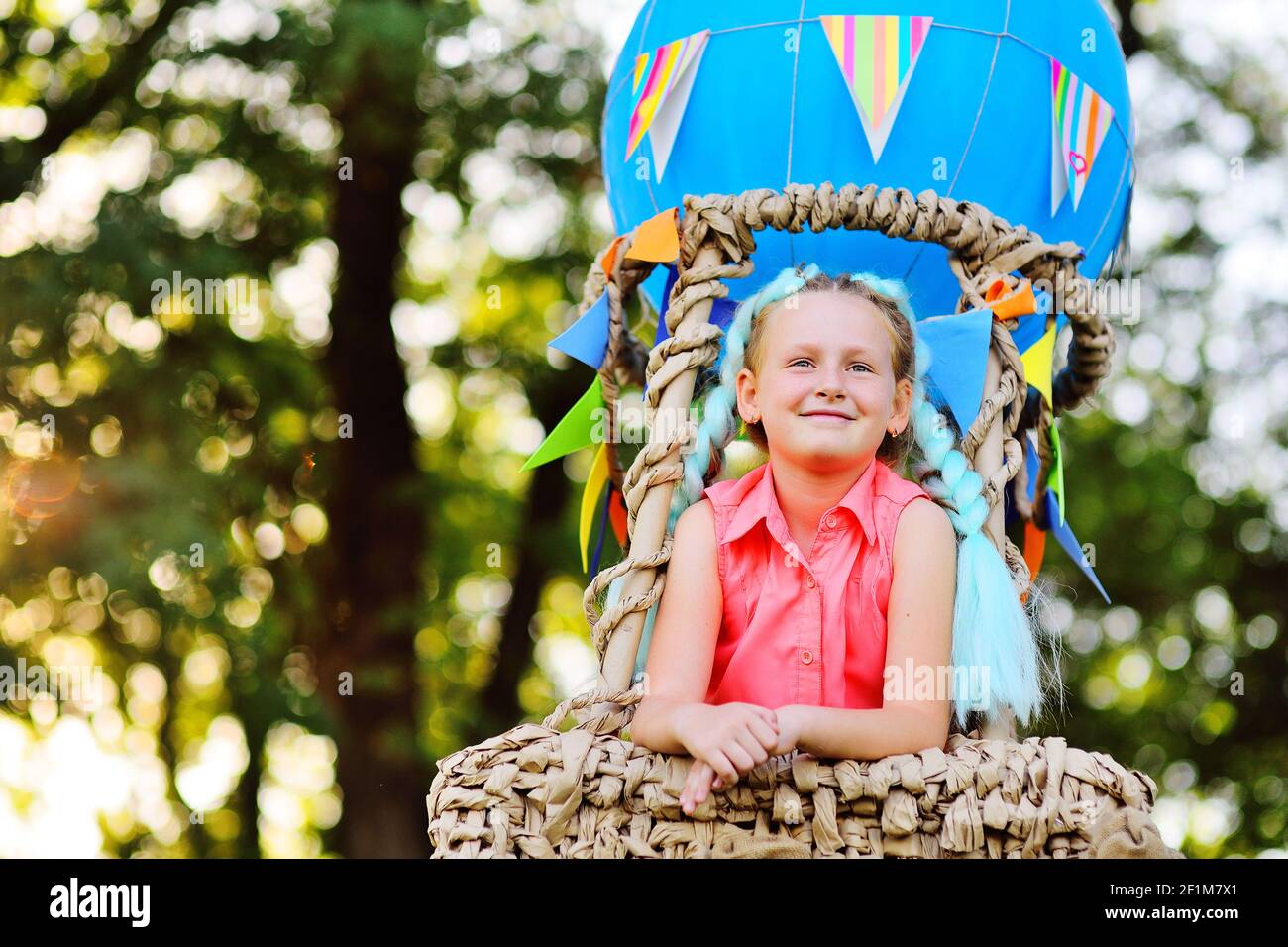 Kleines Mädchen in rosa Kleidung lächelt sitzen in einem Korb mit einem blauen Ballon vor einem Hintergrund von Sonnenlicht und Grün. Stockfoto