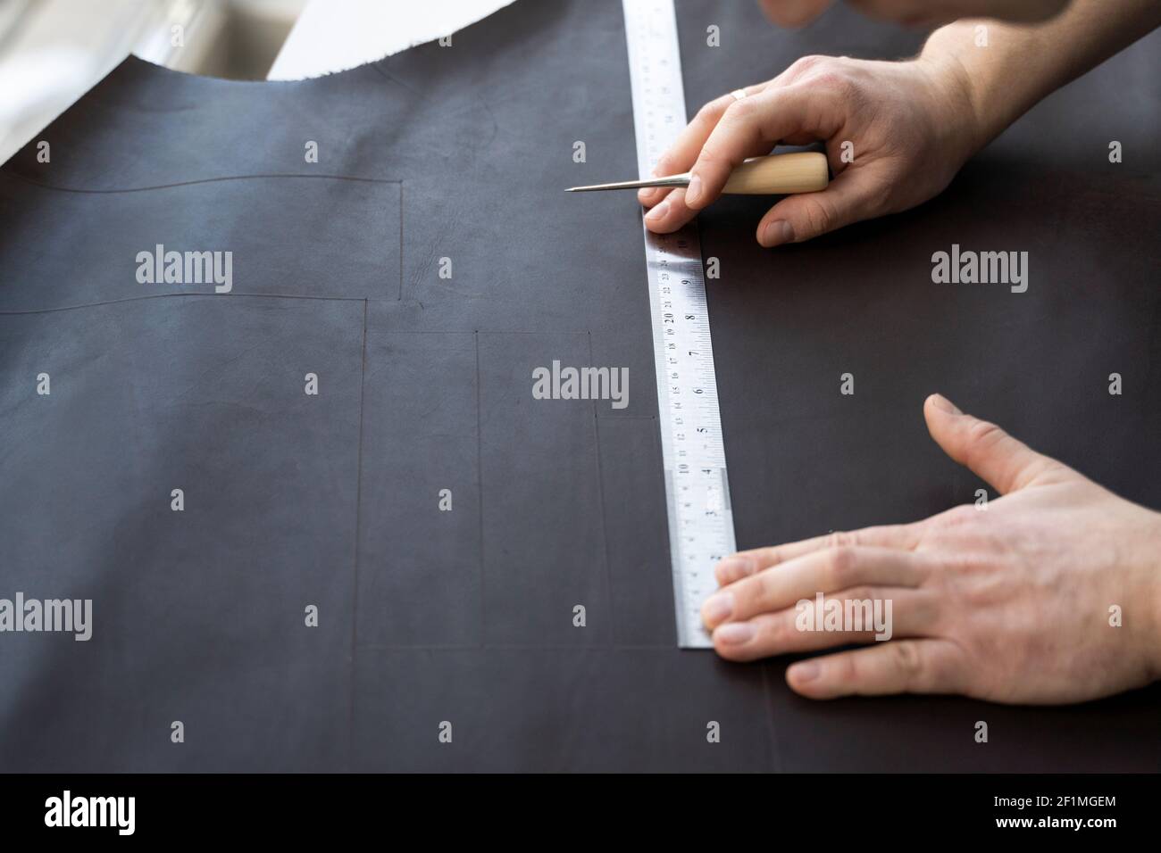 Herrenhand, die eine Ahle und ein Metalllineal hält und an einer Ledertasche in seiner Werkstatt arbeitet. Arbeitsprozess mit einem braunen Naturleder. Handwerker Stockfoto