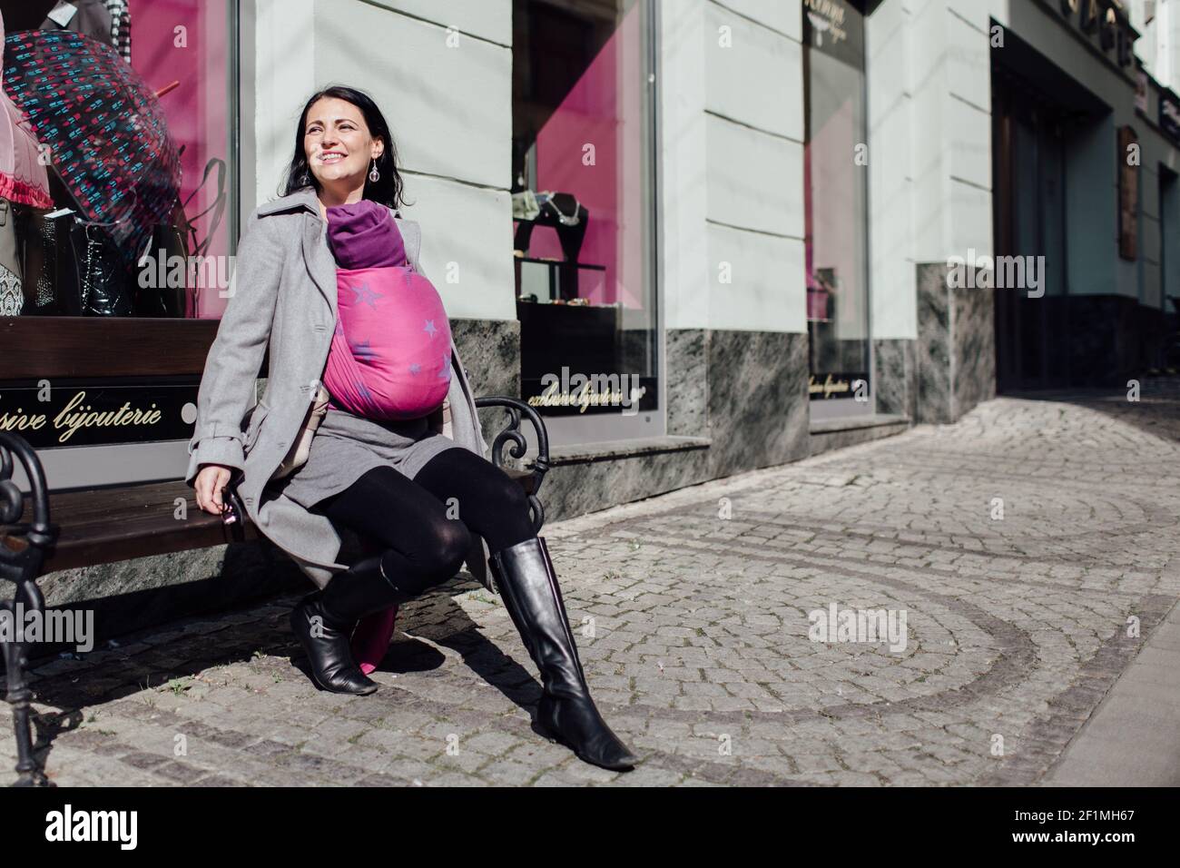 Portrait von trendigen Babytragemutter sitzt auf der Straße Bank in Die Stadt Stockfoto