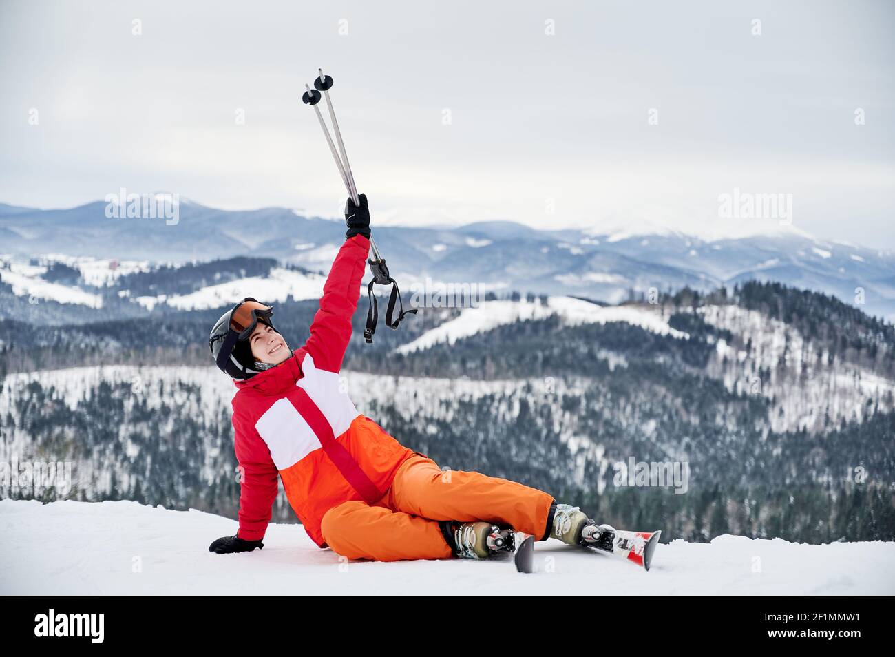 Horizontale Momentaufnahme des Mädchens, das einen lebhaften Skianzug trägt, auf Schnee sitzend, ihre Skistöcke oben haltend. Schräge Berge im Hintergrund. Konzept des aktiven Lebensstils Stockfoto