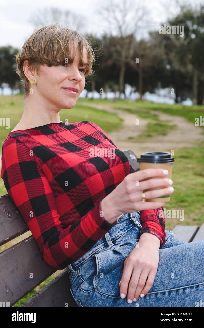 kaukasisch attraktiv hält eine Einweg-Tasse mit Kaffee. Sie trägt Freizeitkleidung und sitzt auf einer Bank in einem grünen Park. Es ist im Frühling Stockfoto