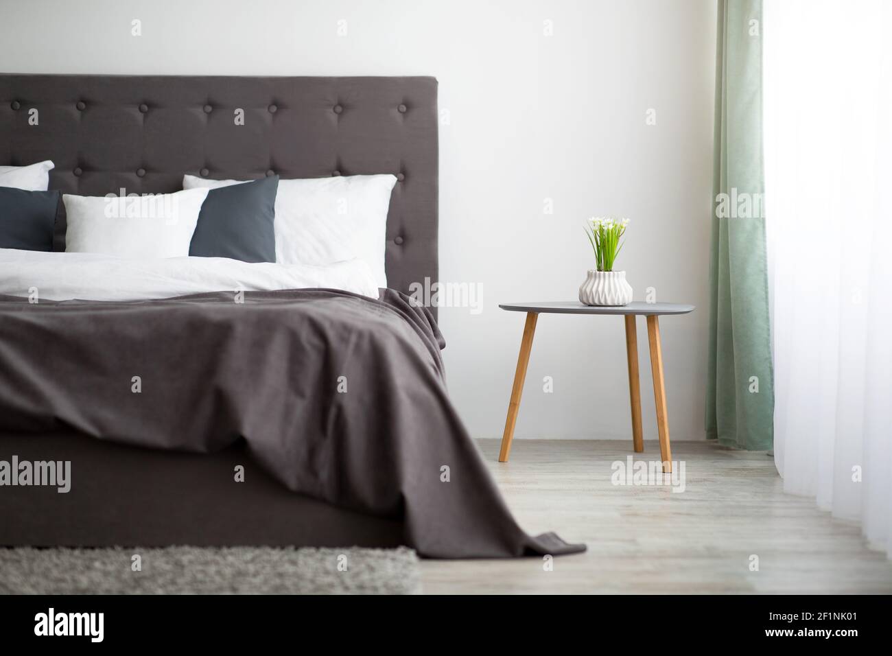 Moderner Stil und modisches Design im Inneren der Wohnung. Bett für zwei Personen mit Decke und Kissen, Topfpflanze auf Tisch auf grauem Wandhintergrund, groß Stockfoto