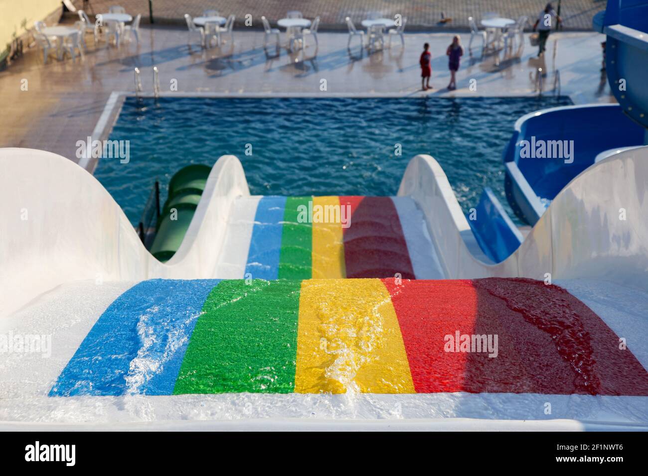 Blick von oben von einer Pool-Rutsche in Regenbogenfarben mit fließendem Wasser. Neben dem blau türkisfarbenen Pool gibt es Tisch und Stühle eines Aquaparks Stockfoto