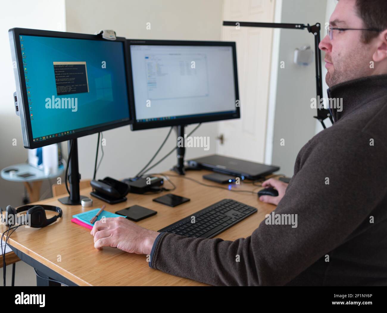 Der Mann sitzt an einem Schreibtisch im Home Office vor zwei Computermonitoren, die auf Augenhöhe auf den Armen angehoben werden. Fernarbeiten. Stockfoto