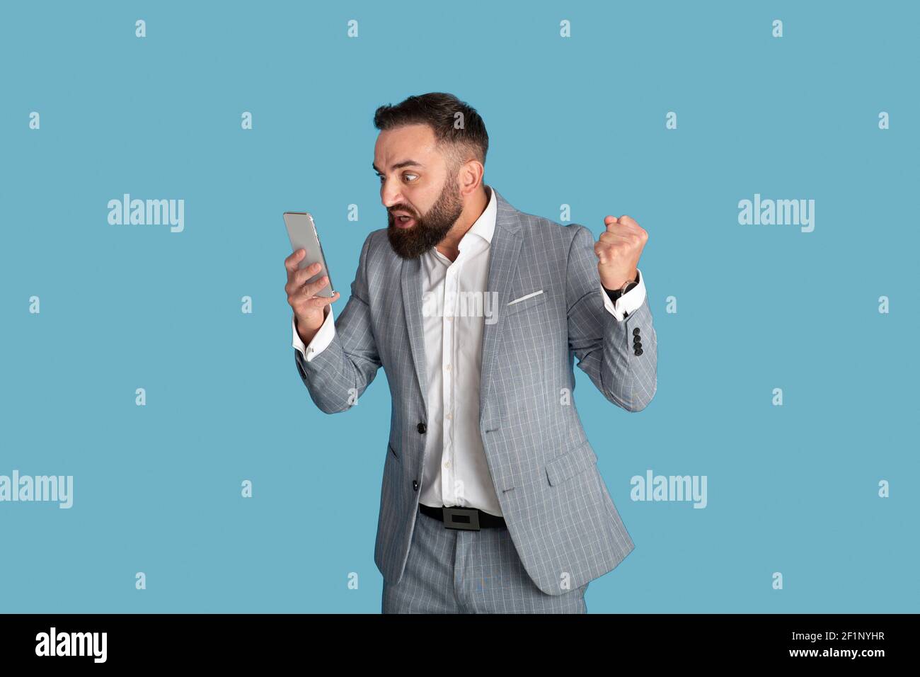 Aggressiver junger Geschäftsmann, der unter Stress am Arbeitsplatz leidet und das Smartphone über blauem Studiohintergrund anruft. Wütender Mitarbeiter im Büro, tausendjährig Stockfoto