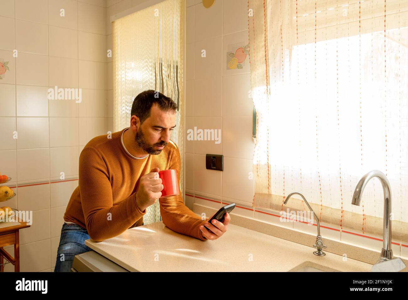 Hübscher bärtiger Mann in einem orangefarbenen Pullover, der das Smartphone in der Küche anschaut, während er eine Tasse Kaffee trinkt. Technologiekonzept Stockfoto