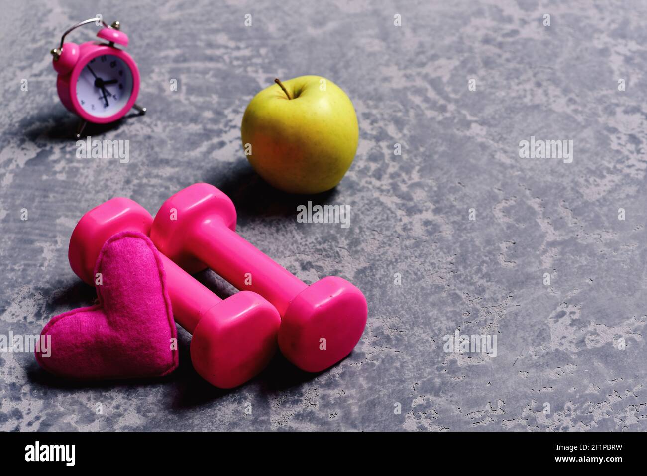 Liebe zum Sport und frühes Training. Damen-Style und Morgen-Workout-Konzept. Hanteln in rosa Farbe neben Apfel, Wecker und weiches Spielzeug Herz auf grauem Hintergrund. Herz, Apfel und Kunststoff-Langhanteln Stockfoto