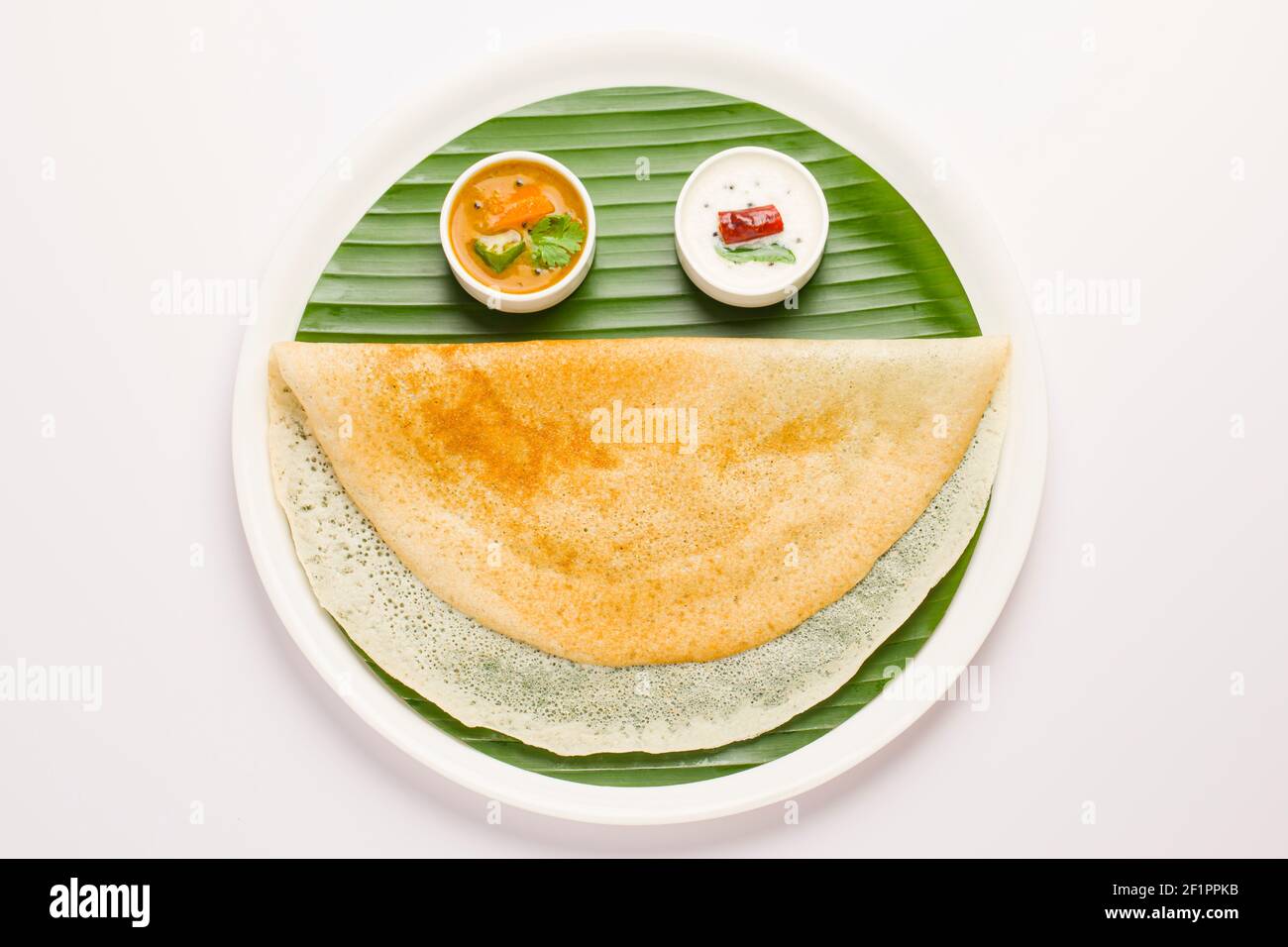 Ghee Roasted Dosa, südindisches Hauptfrühstück, das wunderschön wie ein lächelndes Gesicht auf einem weißen Teller mit Bananenblatt auf weißem Ba angeordnet ist Stockfoto