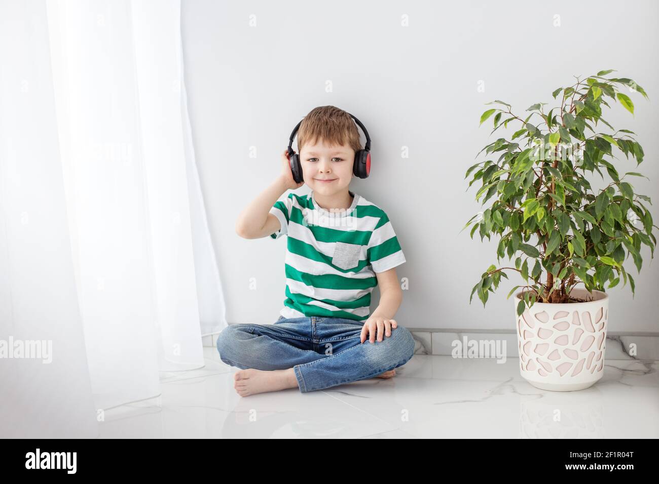 Ein kleiner Junge sitzt auf dem Boden in einem hellen Raum und hört Musik mit Kopfhörern. Stockfoto