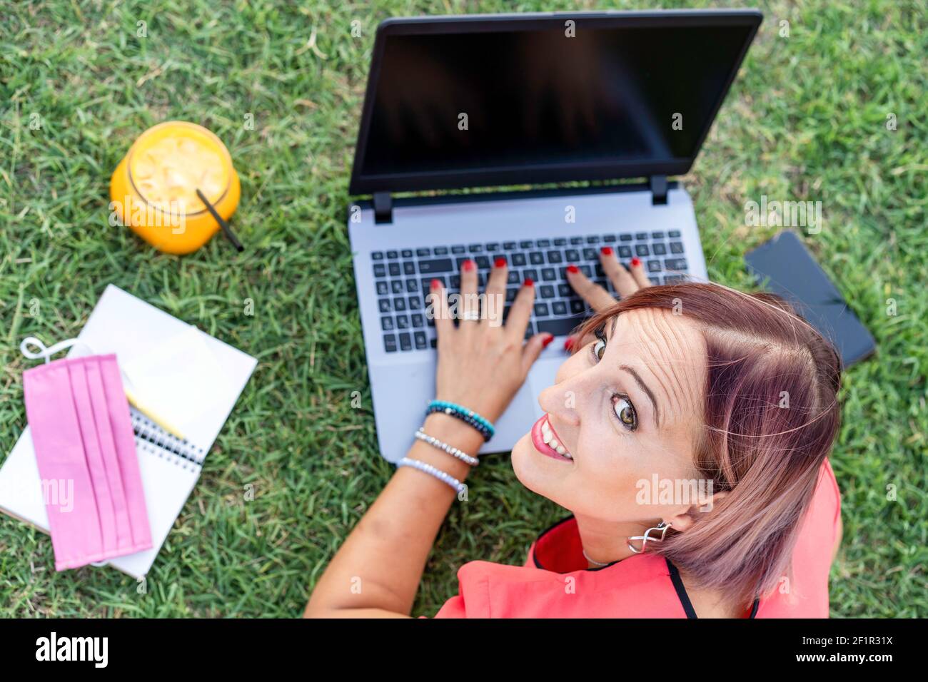 Frau 40 Jahre alt arbeiten im Freien mit Laptop während der Herstellung Gesundes Frühstück - lächelnde Unternehmerin oder Influencerin, die sich hinlegt Auf Rasen tippen o Stockfoto