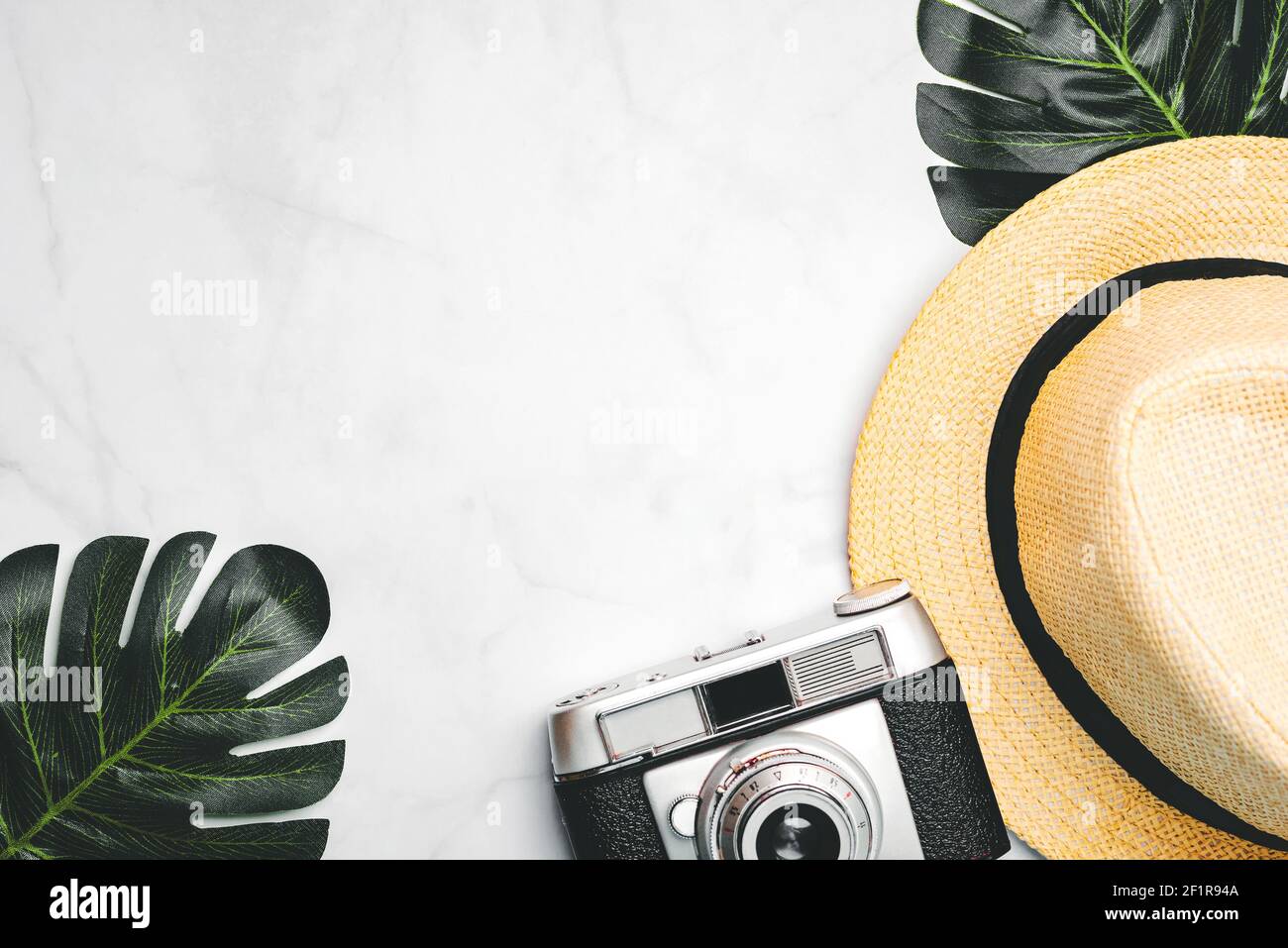 Strohhut mit grünen tropischen Blättern und Vintage-Fotokamera Mit Platz für Text auf weißem Marmorhintergrund.Urlaubsreisekonzept Stockfoto