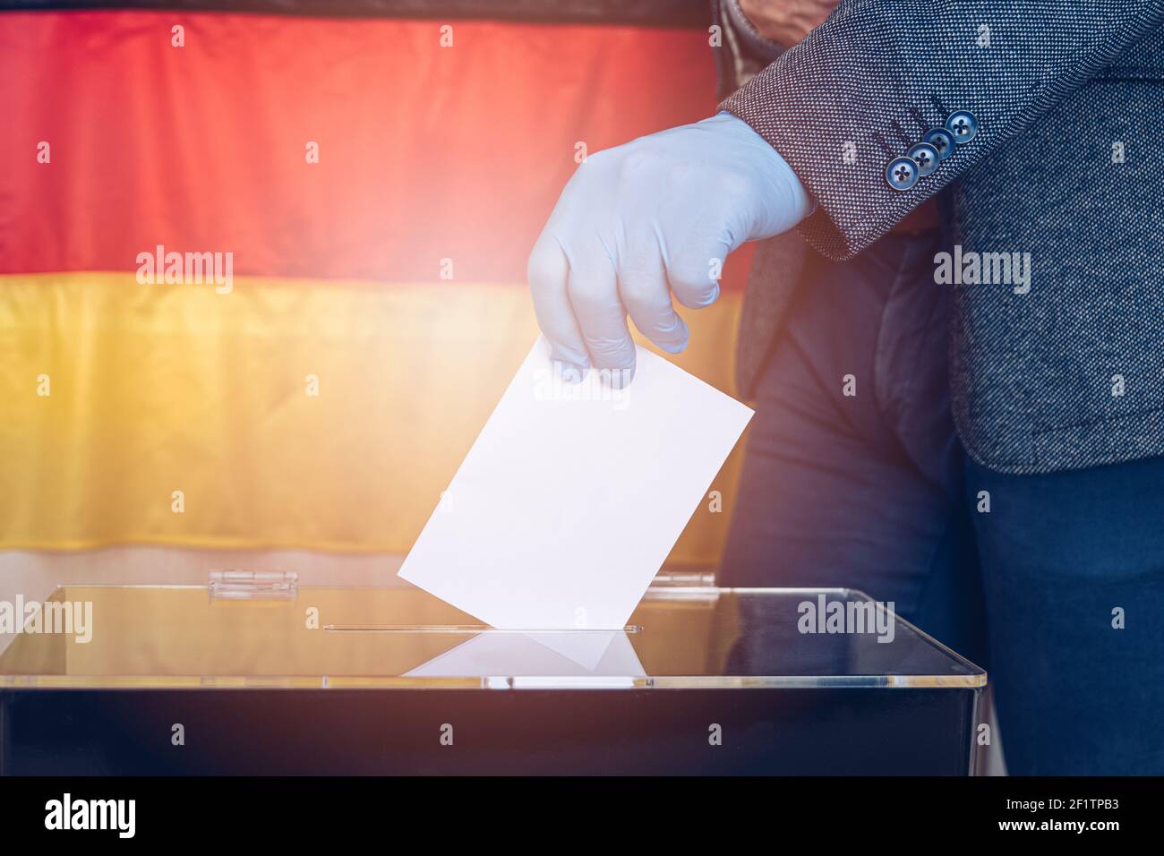 Seitenansicht Nahaufnahme der Hand des Mannes mit Schutzhandschuh, der die Abstimmung gegen den Hintergrund der deutschen Flagge in die Wahlurne setzt. Stockfoto