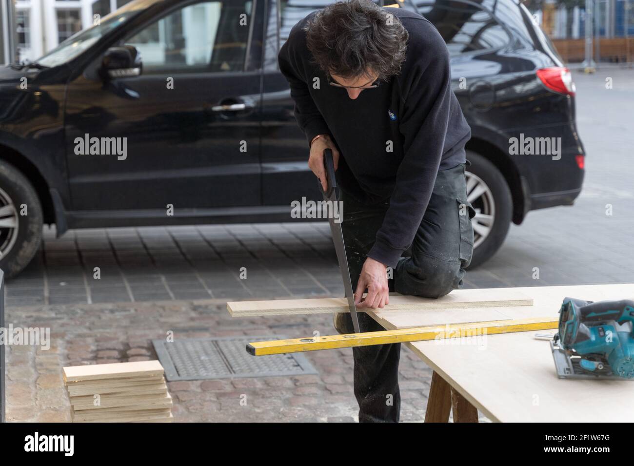 Zimmermann mit Gläsern verwendet Handsäge Querschneiden eine Planke von Sperrholz auf der Arbeitsplatte neben seinem Auto Stockfoto